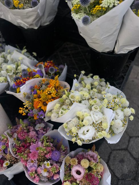 A display of fresh floral arrangements featuring a variety of vibrant flowers, including yellow sunflowers, purple thistles, white roses, and small daisy-like blossoms, all wrapped in white paper. The